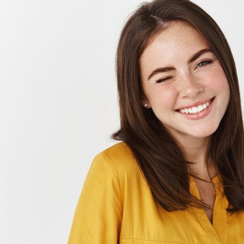Close-up of cute and dreamy young woman smiling, winking at camera to encourage client, standing over white background.