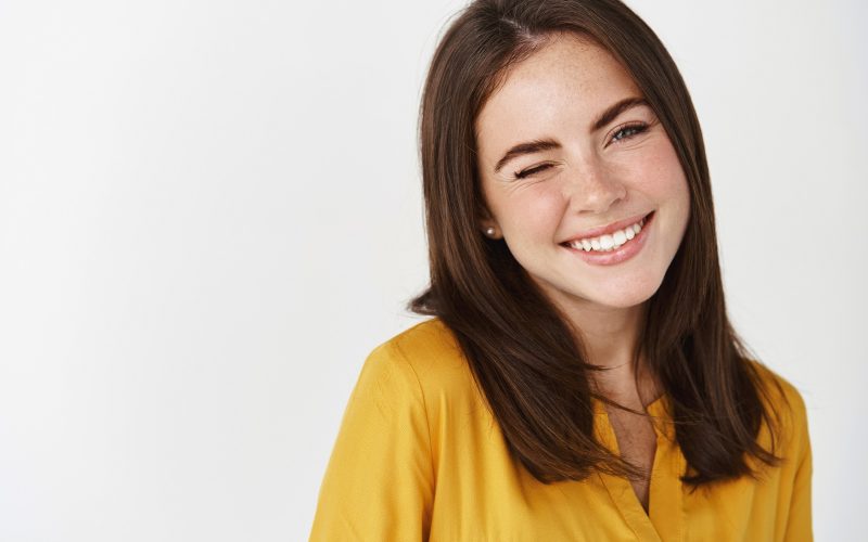Close-up of cute and dreamy young woman smiling, winking at camera to encourage client, standing over white background.